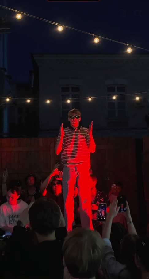 Cheba Man AI example: a man in sunglasses and a striped polo dances on a red-lit stage at a night party while the crowd films